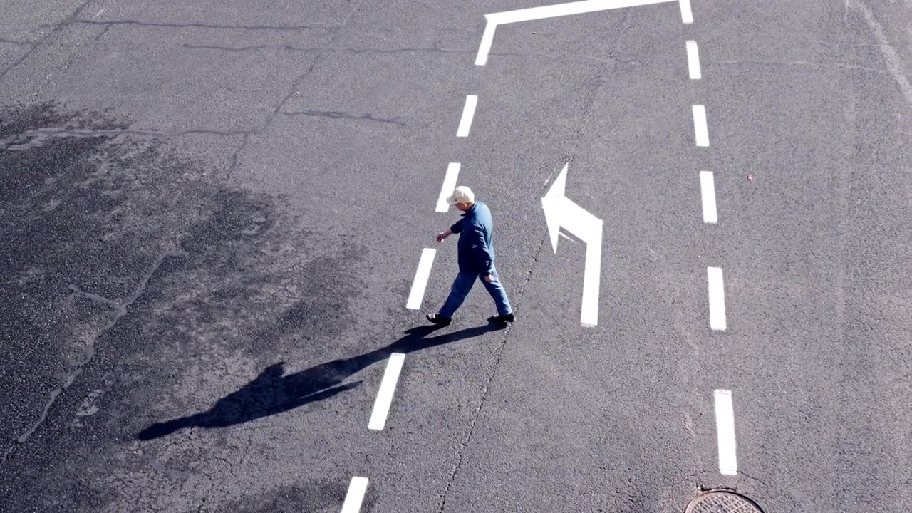 A lone individual walks along a city street with prominent road markings. The scene captures a sense of isolation and movement. Suitable for urban lifestyle or introspective themes.