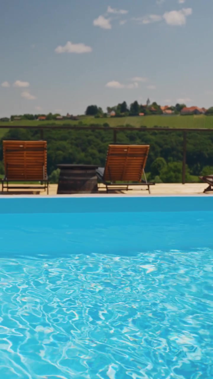 Fredfyldt vertikal video af en klar blå pool, tomme liggestole og frodigt landskab under en solrig himmel. Perfekt til sociale medier og mobilbrug.