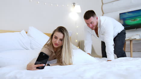 beautiful blonde woman lies on the bed taking selfie. bearded man lies beside looking at telephone camera.