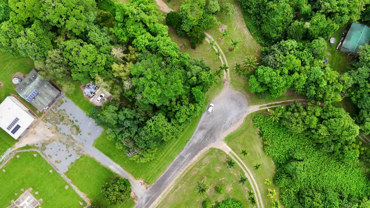 Drone smoothly flies above lush rainforest road intersection, revealing green landscape, buildings, and clearings