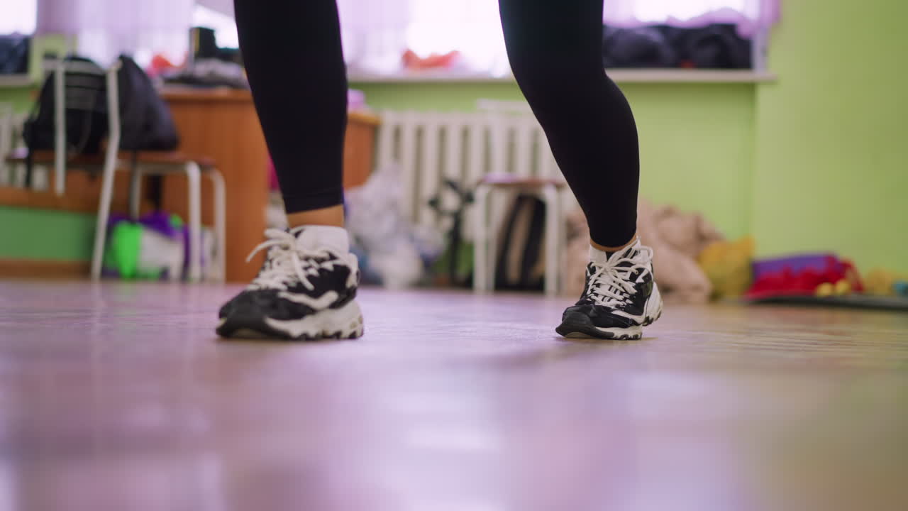 Leg view of girl in black leggings and canvas sneakers bouncing on polished wooden floor during indoor workout, highlighting energy, rhythm, and athletic movement against blurred background setting