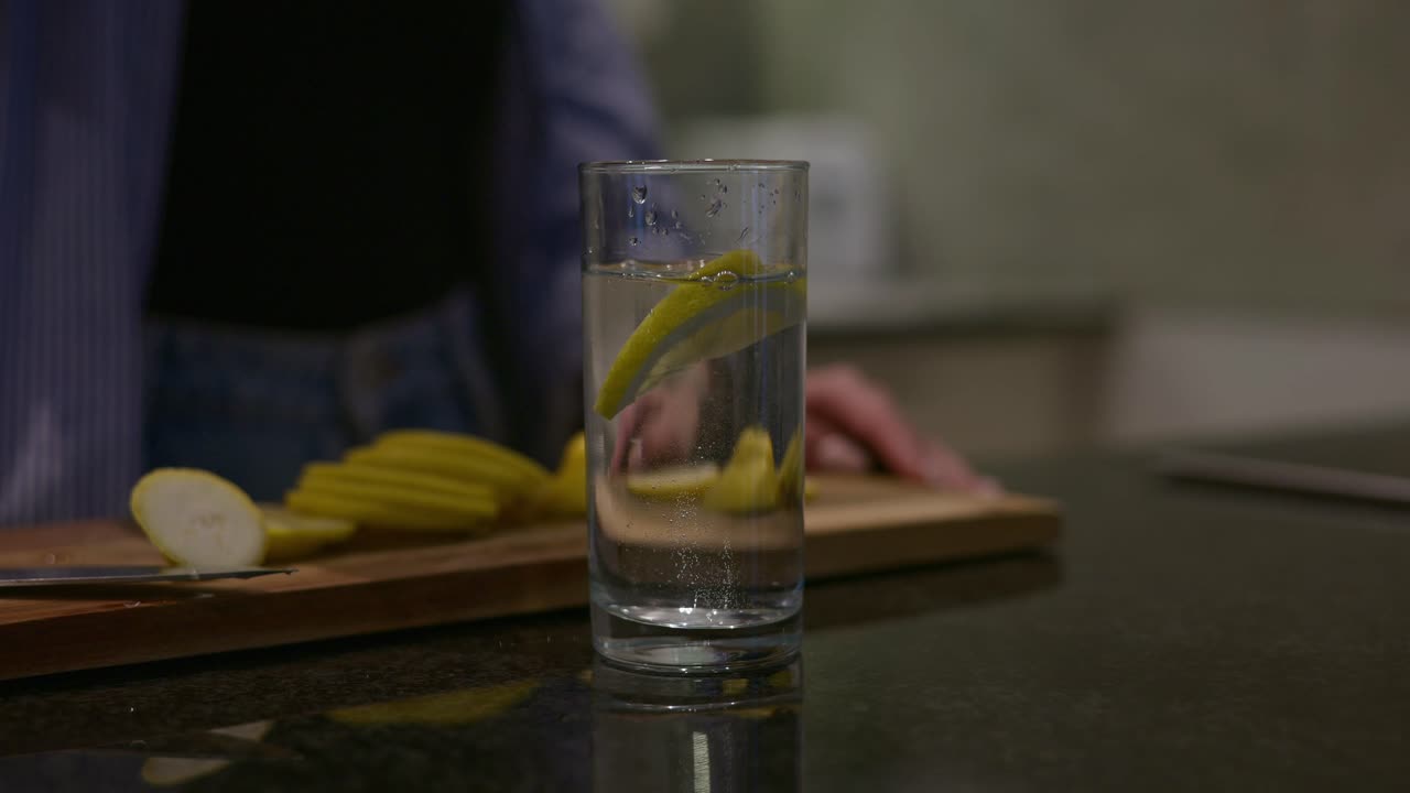 Primo piano di un bicchiere alto di acqua e limone posizionato su un bancone della cucina. Fette di limone fresche e un tagliere sullo sfondo evidenziano la preparazione di una bevanda sana e rinfrescante a casa.