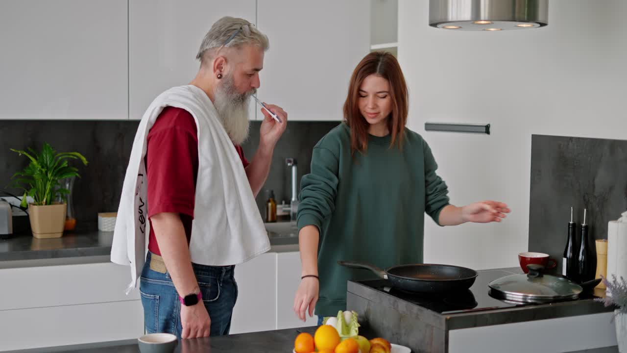 An elderly man with gray hair and a lush beard in a red T-shirt brushes his teeth while a brunette girl in a green jacket makes breakfast and breaks chicken eggs for scrambled eggs in the kitchen in a modern apartment
