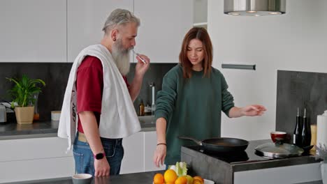 An-elderly-man-with-gray-hair-and-a-lush-beard-in-a-red-T-shirt-brushes-his-teeth-while-a-brunette-girl-in-a-green-jacket-makes-breakfast-and-breaks-chicken-eggs-for-scrambled-eggs-in-the-kitchen-in-a-modern-apartment