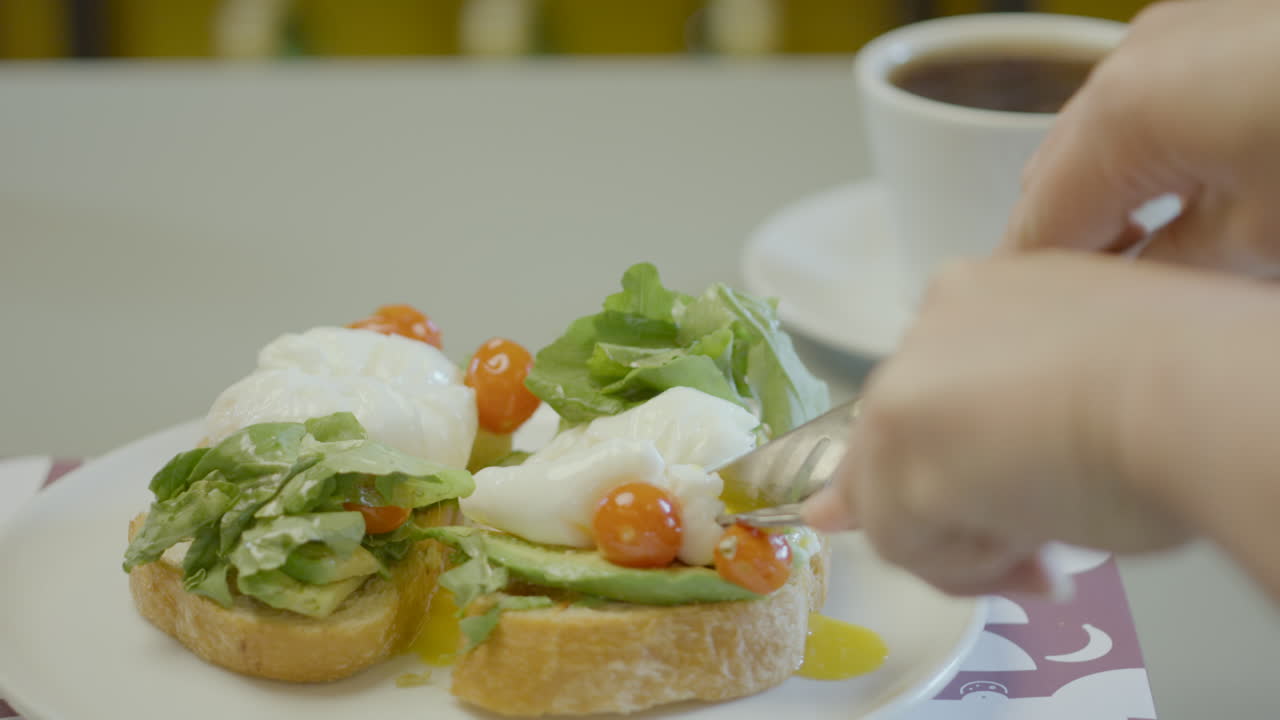 Close-up of a plate with rustic bread toast topped with avocado, lettuce, cherry tomatoes and poached eggs, next to a cup of hot coffee