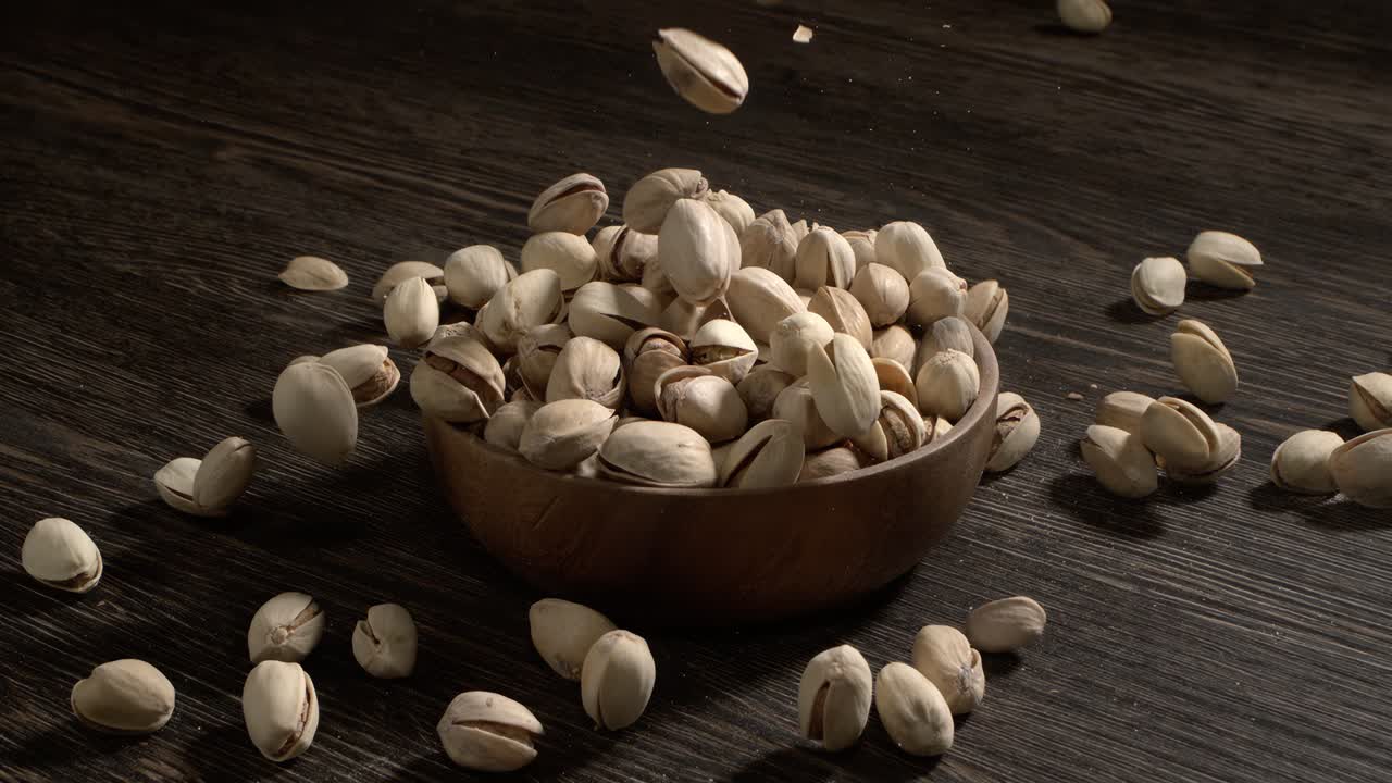Ciotola di legno piena di pistacchi su tavolo di legno scuro, gustosa frutta secca salata sparsa intorno, concetto di snack salutare e cibo naturale. Scatto con telecamera super slow motion a 1600 fps