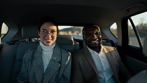 Portrait-of-a-happy-brunette-girl-in-round-glasses-a-businesswoman-in-a-gray-suit-and-her-male-colleague-with-Black-skin-in-a-brown-jacket-during-their-trip-in-a-modern-car-in-a-salon-outside-the-city