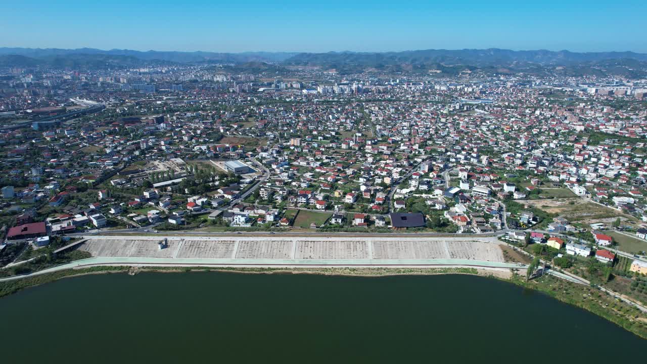 Tirana outskirts appears behind the dam reservoir with urban expansion and crowded neighborhoods, demographic challenges