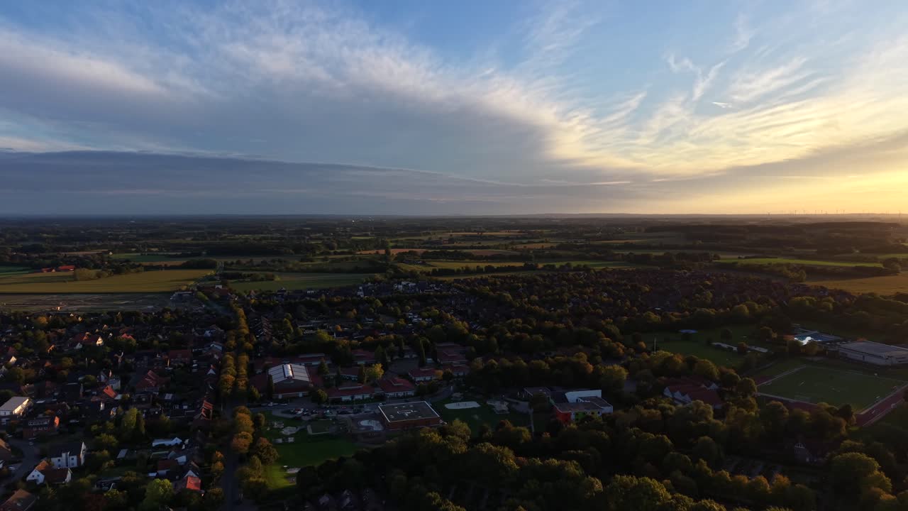 Cloudy golden fall evening in German village. Aerial wide shot. Colored trees and fields in late summer. Houses and Homes in peaceful town. Soccer field stadium of city. Sunset time