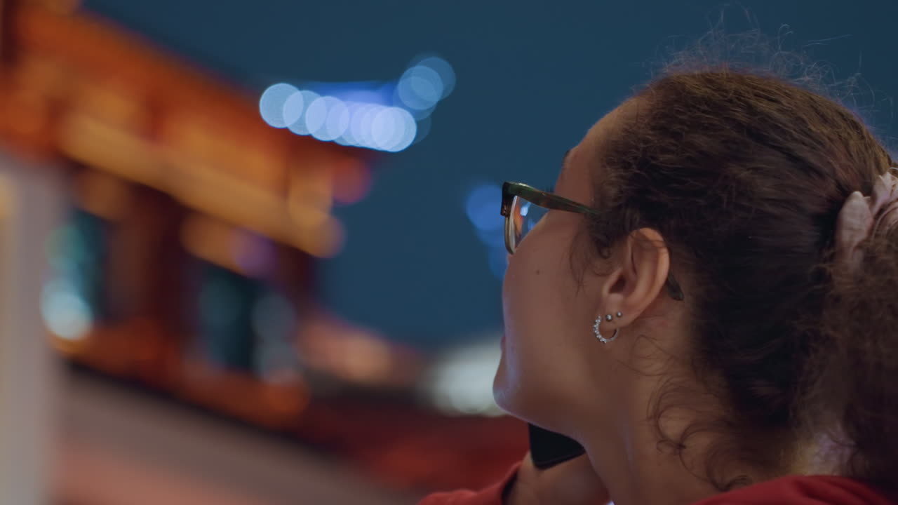 Telefonata notturna in città, giovane donna con felpa rossa con cappuccio che guarda in alto contro la facciata illuminata del tempio, primi piani spontanei catturano espressioni riflessive durante la conversazione, luci bokeh soffuse, occhiali, capelli ricci.