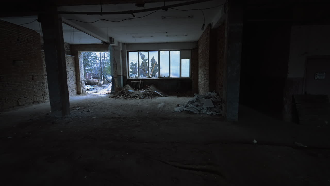 Approaching the large broken window and the big doorway in the spacious premise. Piles of debris are at the walls. Snow falls outdoors.