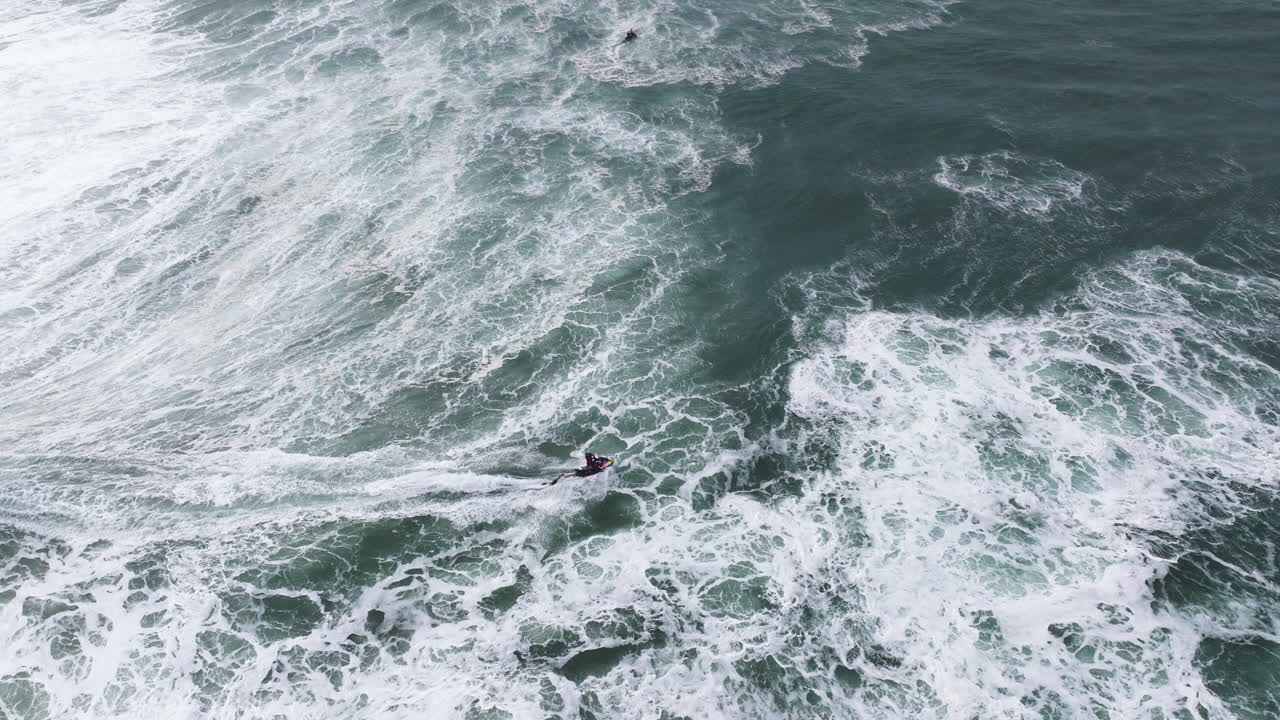 Aerial drone shot of big wave surfer holding onto back of a jet ski in Nazare, Portugal, Europe. Nazaré, big wave surfing town. Shot in ProRes 422 HQ