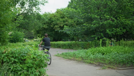 back view of a boy wearing grey trouser and black top riding his bicycle along a paved road surrounded by lush greenery and trees, with a blurred view of cars moving in the background