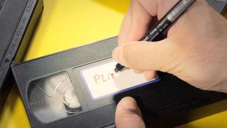 writing the text play me on the white label of an old vhs videotape , with a marker