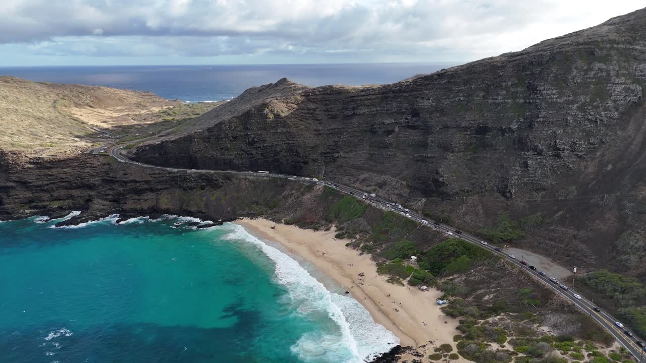 Drone aerial view of a scenic coastal road on Oahu, Hawaii. Winding along lush tropical mountains with ocean views, waves, and dramatic island coastline