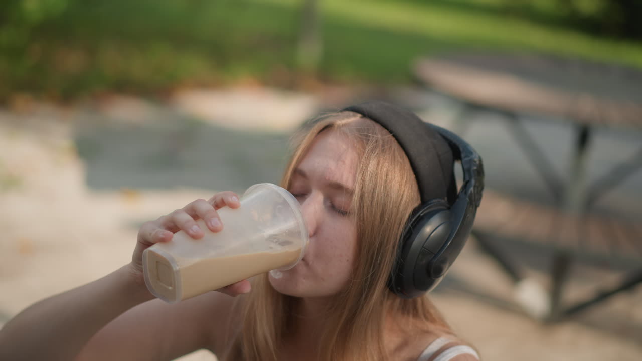 jovem com fones de ouvido tomando café gelado, vibe relaxada de dançarina de rua em um banco de parque, gorro e blusa sem mangas, calçada com luz solar salpicada, close-ups espontâneos, pausa casual entre os movimentos, sorriso contente