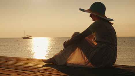 Una-Mujer-Joven-Se-Encuentra-Con-El-Amanecer-En-El-Muelle,-Se-Sienta-Y-Mira-El-Sol-Y-El-Barco-En-El-Mar-Dre