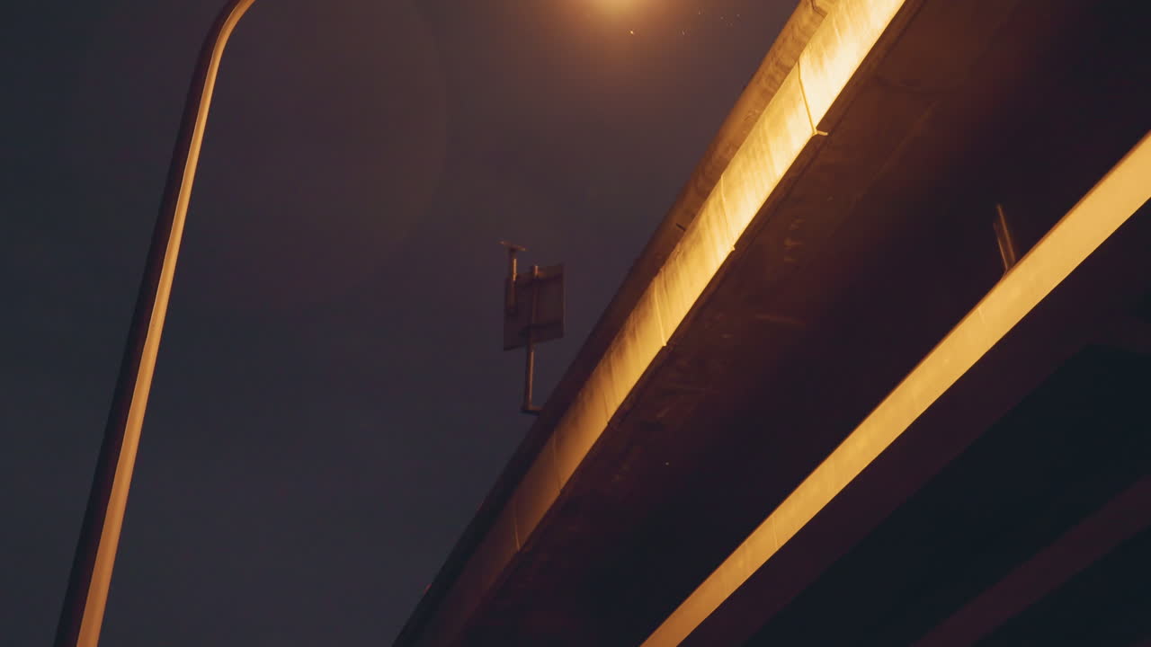 Night overpass illuminated by lamp, golden glow on concrete underside of bridge, empty urban scene with distant surveillance fixture, cinematic shadow and quiet industrial atmosphere
