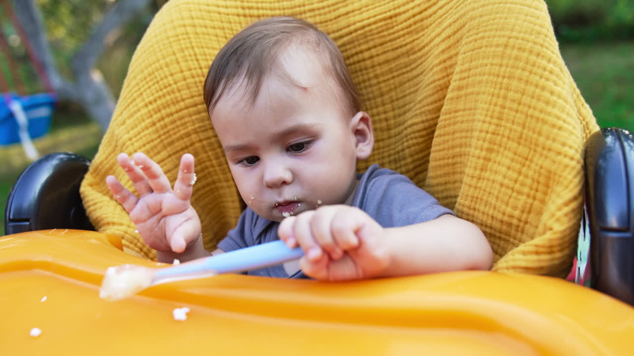 Caucasian baby boy sitting in yellow feeding chair. Little kid with smudged face after feeding turns spoon in hands and looking at it intently. Close up.