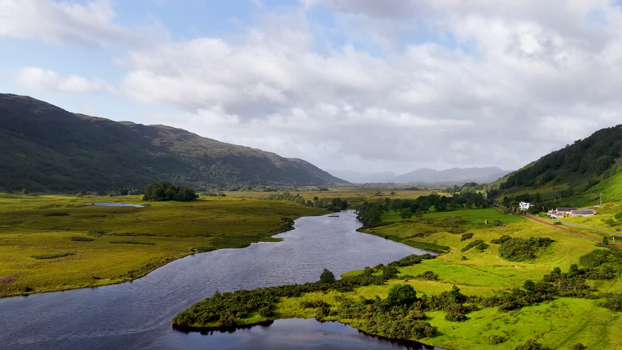 Drone glides over lush river valley, green meadows, distant mountains, under soft daylight clouds