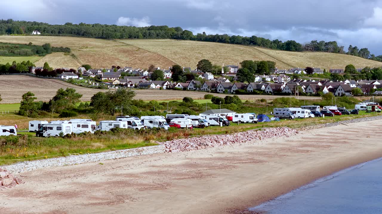 Wide shot pans across beachfront caravans, coastal village, and rolling hills under bright daylight