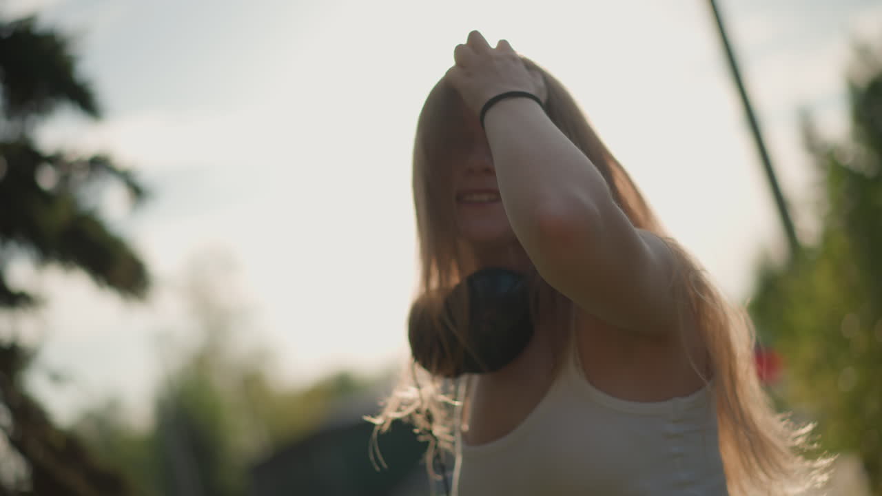 garota sorrindo com fones de ouvido ao pôr do sol, cabelo iluminado por trás esvoaçante, reflexo de lente radiante e risada espontânea em retrato ao ar livre na hora dourada em rua tranquila com sombras suaves e brilho quente, energia divertida