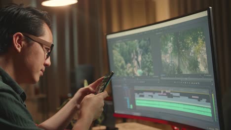 asian male video editor works and using mobile phone with his personal computer, he works late at night