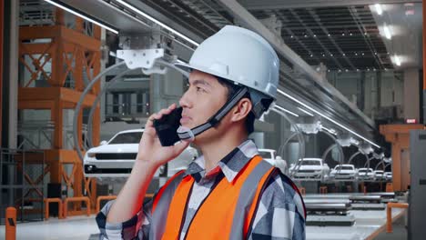 vista lateral de cerca de un ingeniero masculino asiático con casco de seguridad hablando en su teléfono inteligente mientras está de pie en una fábrica de automóviles ev