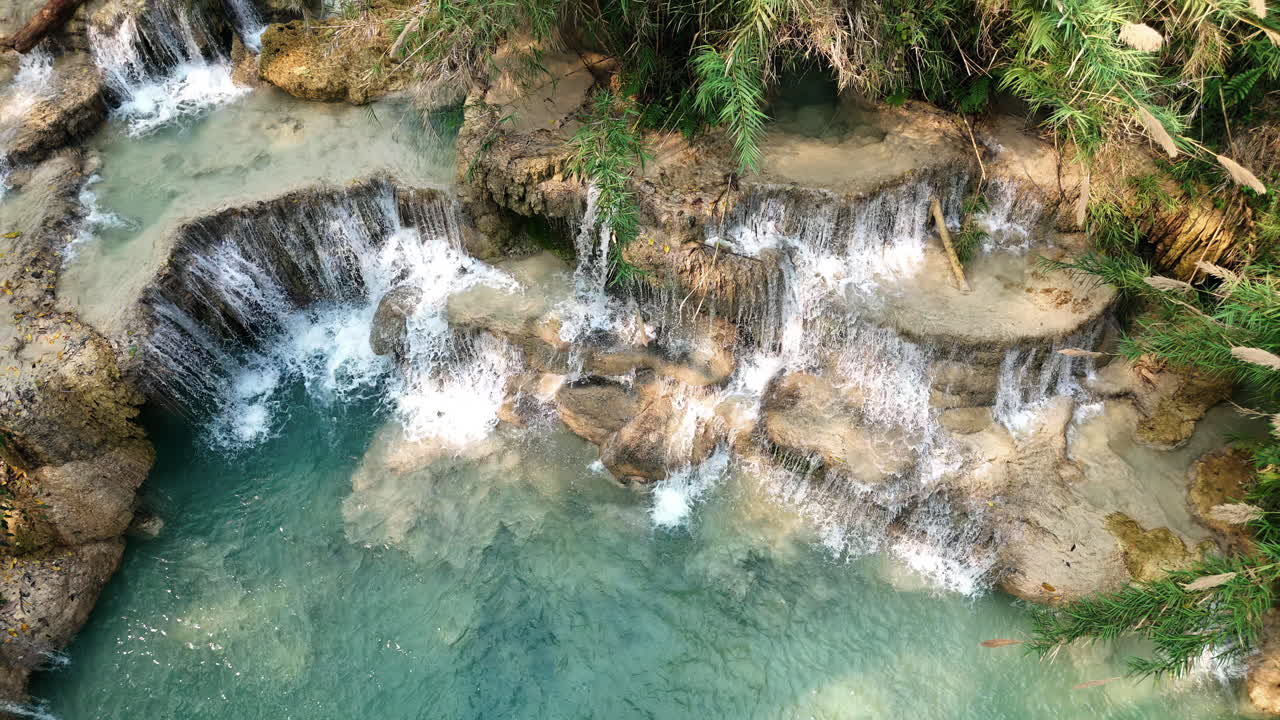 Veduta aerea con drone di piscine turchesi e cascate calcaree a cascata alle Cascate Kuang Si, che scorrono attraverso terrazze rocciose e vegetazione della giungla nel Laos settentrionale