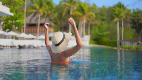 Back-view-of-a-female-tourist-standing-half-submerged-in-a-swimming-pool-in-some-tropical-destination,-rising-her-arms-above-her-head-and-enjoying-her-vacations