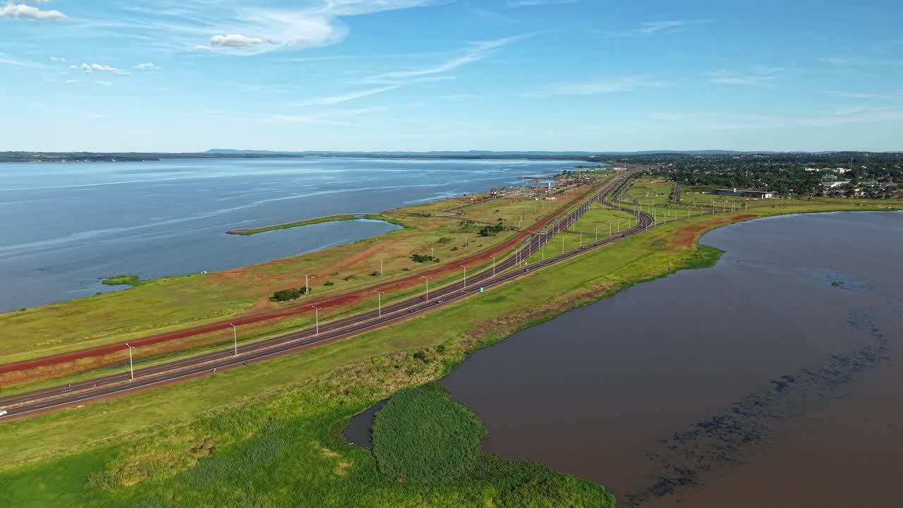 Aerial view of Acceso Sur in Posadas, Misiones, Argentina, shows a highway running along grassy riverbanks beside the Paraná River on a bright summer day