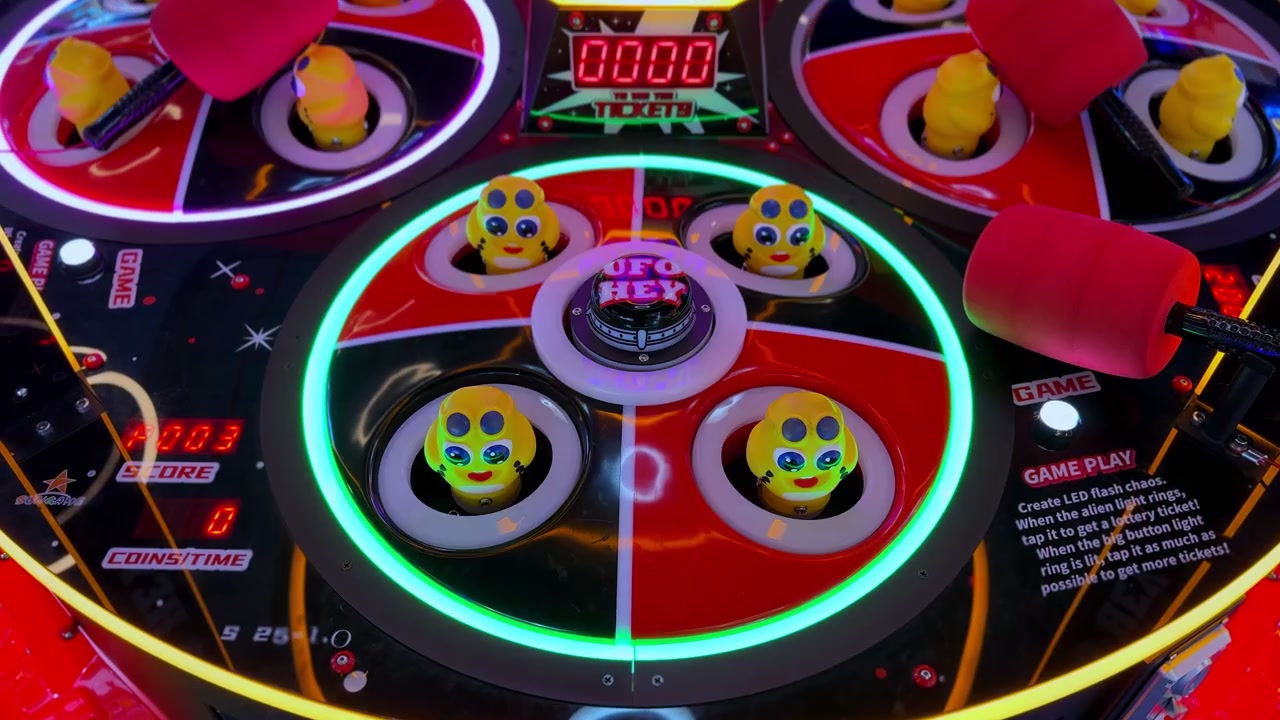 Vista ravvicinata di un colorato gioco arcade in stile "whack-a-mole" al neon, al chiuso. Figure giocattolo aliene gialle spuntano da anelli LED verdi luminosi in attesa di essere colpite da martelli di schiuma rossi per i biglietti premio