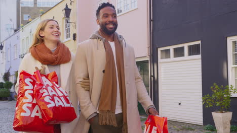 Multi-cultural-couple-arm-in-arm-as-they-walk-along-cobbled-mews-street-on-visit-to-city-in-autumn-or-winter-carrying-sale-shopping-bags---shot-in-slow-motion