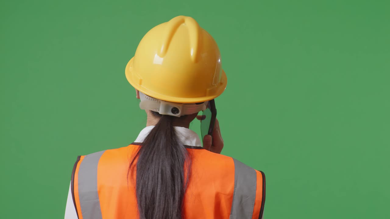 vista de atrás de una ingeniera asiática con casco de seguridad hablando en un teléfono inteligente mientras está de pie en el estudio de fondo de la pantalla verde