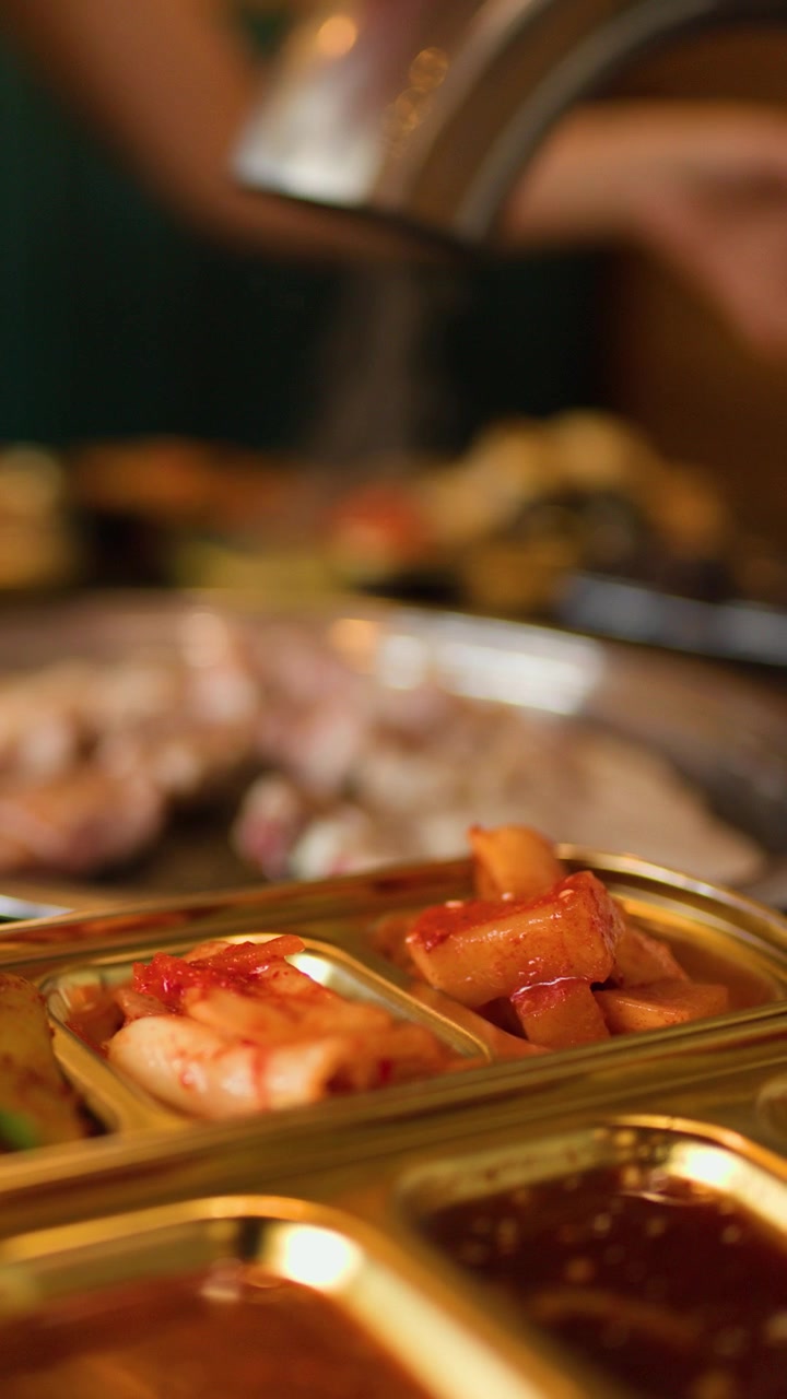 El makasla ızgara et keserken, duman yükseliyor, altın tepsilerde kimchi ve mezeler duruyor