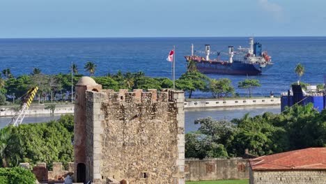 Vista-Aérea-Del-Castillo-De-La-Fortaleza-De-Ozama-Con-Bandera-Ondeante-Y-Barco-Viejo-En-El-Mar-De-Fondo---Santo-Domingo,-República-Dominicana