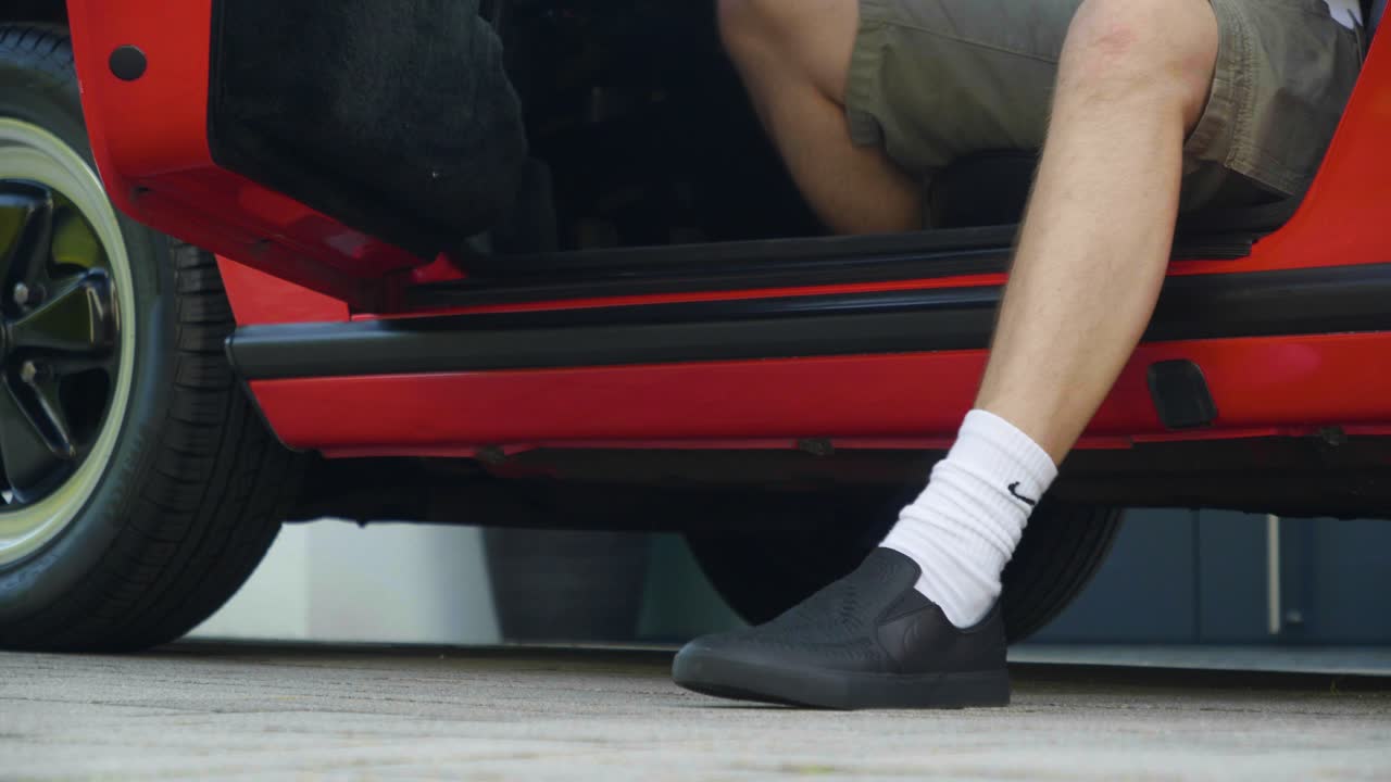 빨간 차에서 반바지, 검은 신발, 흰색 양말을 신고 나온 사람의 낮은 핸드헬드 중간 샷, 카메라가 따라가는 동안 슬로우 모션