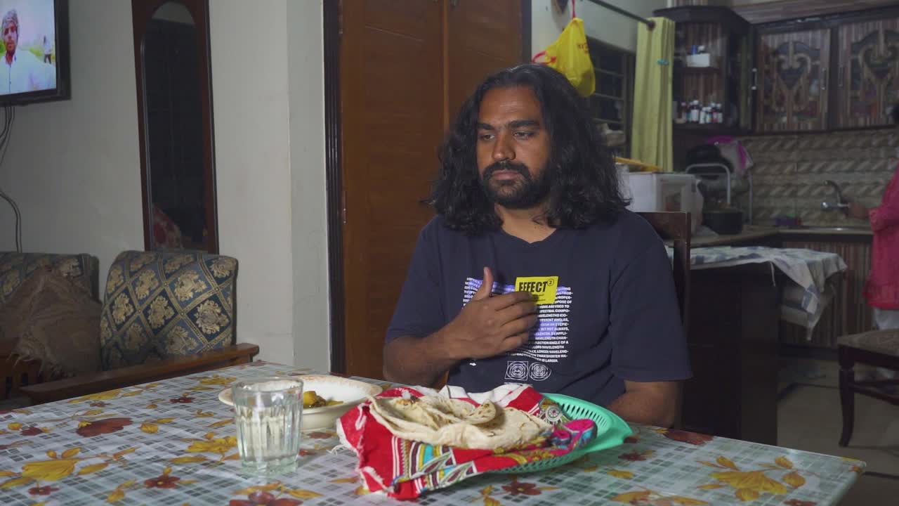 An Indian man thanksgiving to the Lord by making a sign of the trinity at the beginning of his meal at his home's dining room table while his wife works in the kitchen, Slow-mo