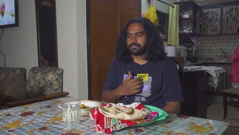 an indian man thanksgiving to the lord by making a sign of the trinity at the beginning of his meal at his home's dining room table while his wife works in the kitchen, slow-mo