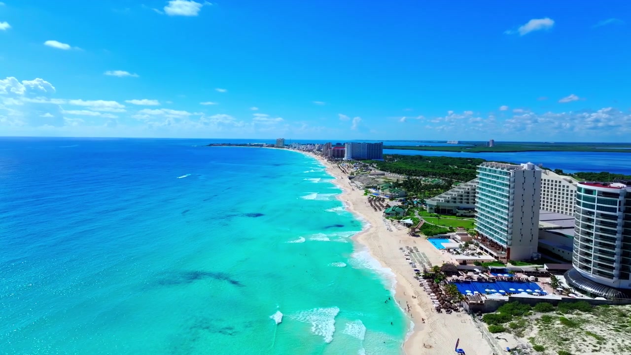 Vista aerea lungo la spiaggia della Zona Hotelera di Cancun, Messico, con l'acqua turchese dei Caraibi che lambisce la riva di sabbia bianca fiancheggiata da resort sotto un cielo luminoso.