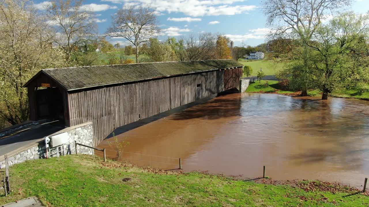 Aerial push in on covered bridge above muddy flooded river on beautiful autumn day