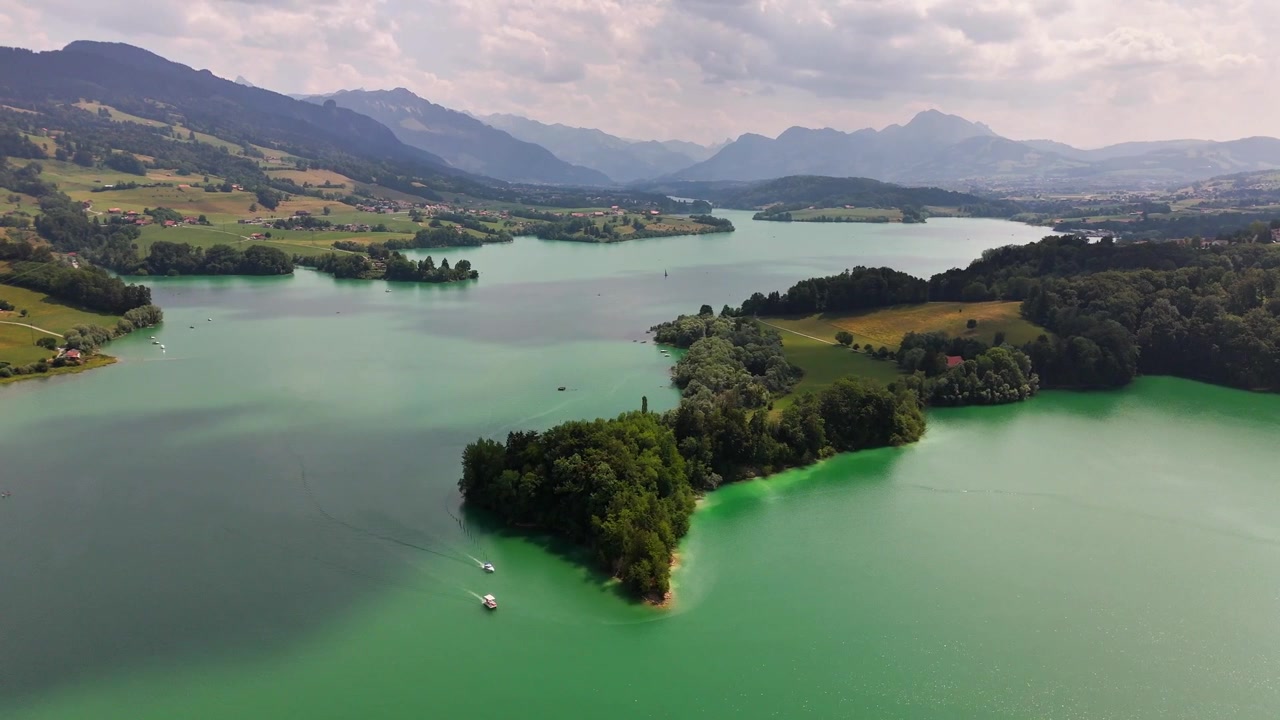 Eine Drohne fliegt über den Greyerzersee bei Pont-la-Ville, Schweiz, und zeigt eine bewaldete Halbinsel, türkisfarbene Buchten, kleine Boote, Ackerland und Bergkämme unter einem bewölkten Himmel.