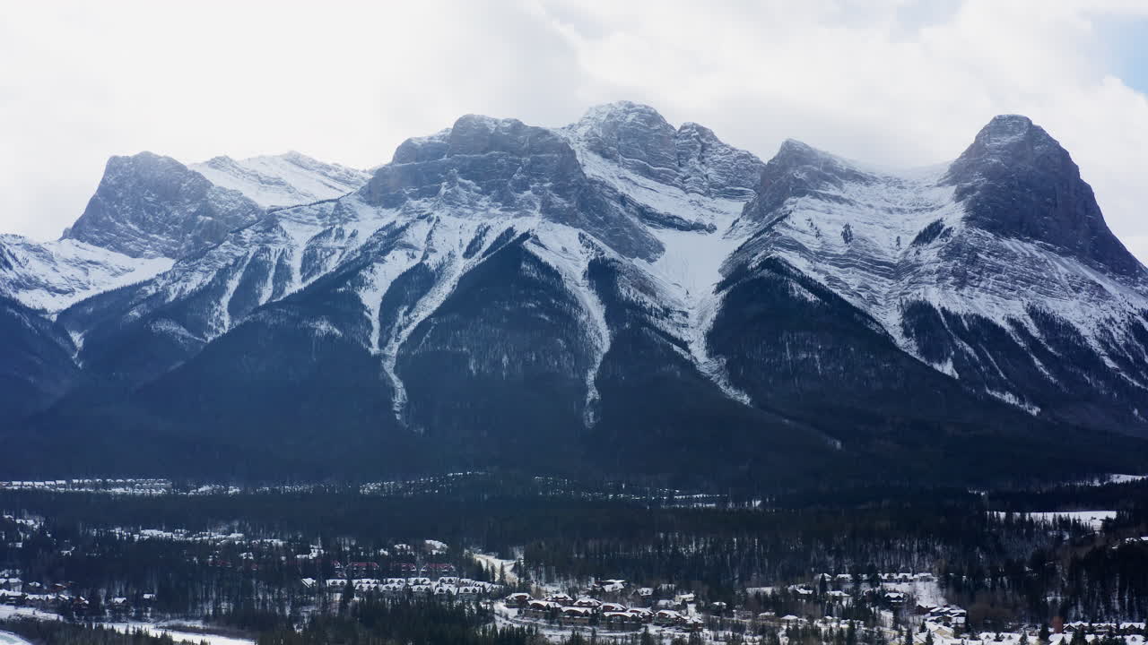Drammatica panoramica con drone rivela le imponenti vette delle Tre Sorelle sopra una tranquilla città di Canmore, immersa nella foresta innevata delle Montagne Rocciose canadesi