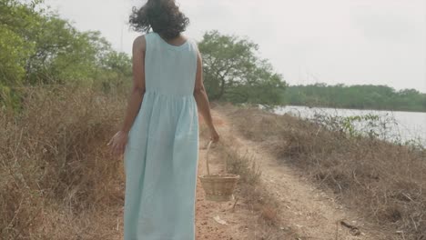 back shot of a woman in a blue dress walking in close to the river with a basket in her hand