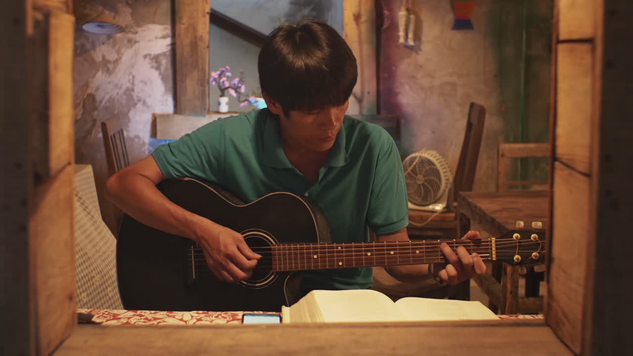 Guardando attraverso una finestra aperta, un giovane uomo asiatico suona la chitarra seduto su una sedia in una stanza rustica.