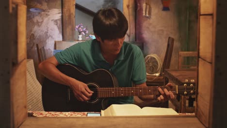 looking through an open window at a young asian male playing a guitar while on a chair in a rustic room