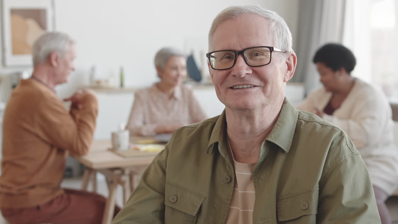 Tilting up POV of grey-haired Caucasian man smiling, wearing glasses, talking and looking on camera, sitting in wheelchair in foreground of blurred people