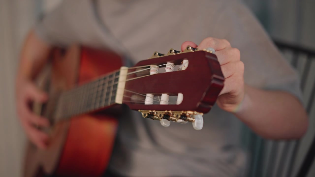 Closeup acoustic guitar headstock and hands tuning during home lesson young student practicing chord progressions on warm wooden guitar, seated on chair in casual room, focused fingers, remote tutor