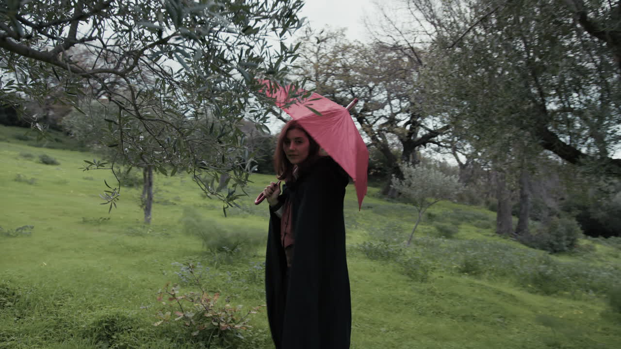 una mujer joven camina por el prado entre los árboles con un paraguas rojo en un día lluvioso