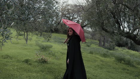 young woman walks on the meadow among trees with a red umbrella on a rainy day